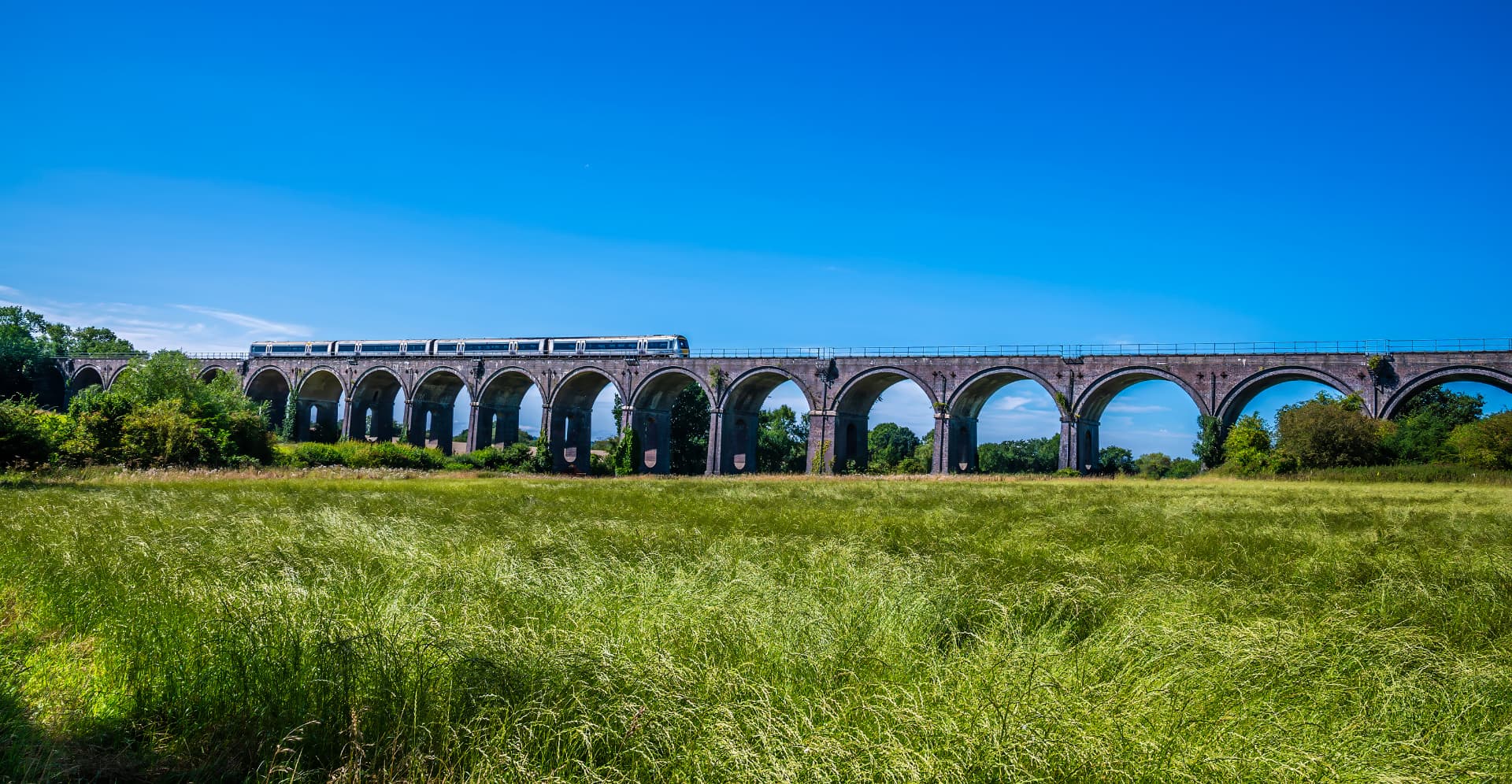 Passenger train crossing a brick railway viaduct above a green meadow